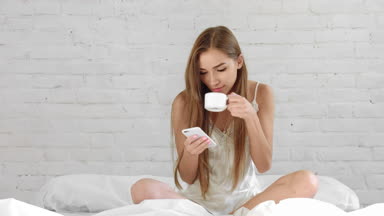Attractive woman sitting in bed, drink coffee and chatting online on cell phone