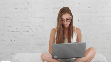 Girl in sleepwear and glasses sitting on the bed and messaging via laptop