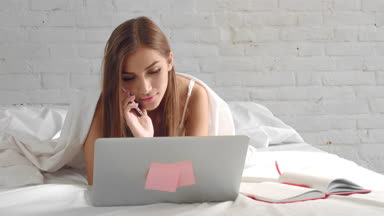Young woman working on laptop and speaking phone in bedroom