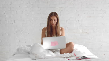 Young attractive female writer sitting in bed and writing book in laptop