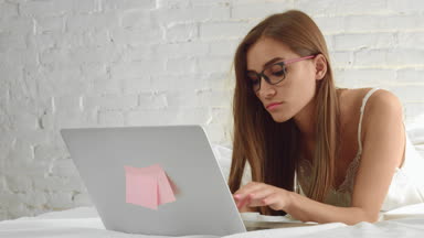 Woman in glasses lying in bed and working on laptop