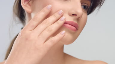 Woman gently touches her face with her fingers on light grey bg