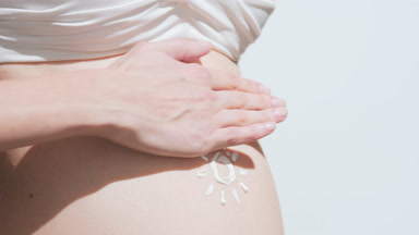 Pregnant woman in a white bra applying sunscreen on her belly