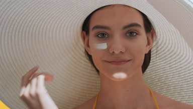 Woman in a big white hat applies spf cream on her cheek smiling
