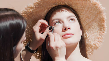 Woman puts makeup on female model in a straw hat using eyeliner