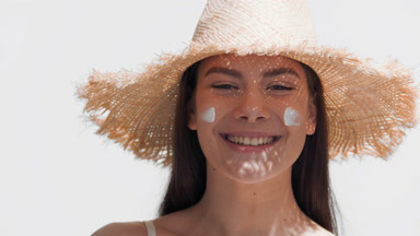 Girl in a straw hat puts spf cream on her cheeks and smiles wide
