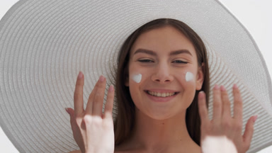 Woman in a white hat applies sunscreen on cheeks and smiles wide