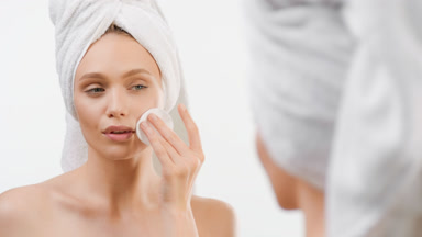 Woman touches face with cotton pad smiling in front of mirror