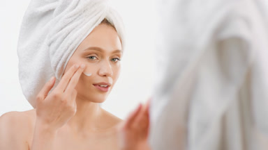 Woman applies face cream on cheek smiling in front of mirror