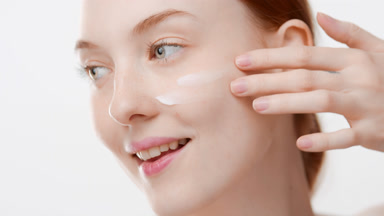 Girl with long orange hair applies moisturizer on cheek smiling