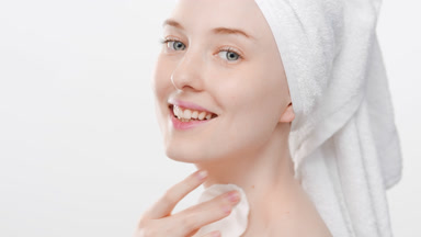 Caucasian woman with head towel uses cotton pad for neck smiling