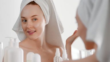 Young woman puts skin care cream on cheek in front of the mirror
