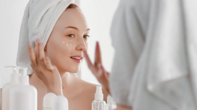 Pretty Caucasian girl applies moisturizer on cheek smiling wide
