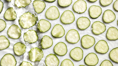 Poured water makes ripples over cucumber slices on white bg