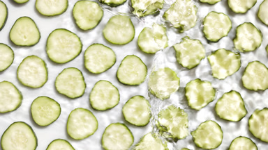 Poured water makes splashes over cucumber slices on white bg