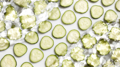 Poured water splashing over cucumber slices on white bg