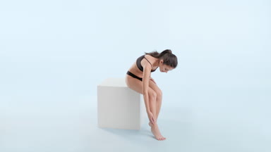 Woman strokes legs sitting on cube and looks at camera smiling