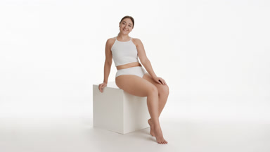 Young plus size woman strokes leg sitting on white cube platform