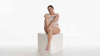 Plus size woman strokes arm sitting on cube platform and smiling