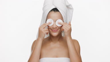 Woman with towel on hair covers eyes with cotton pads and smiles
