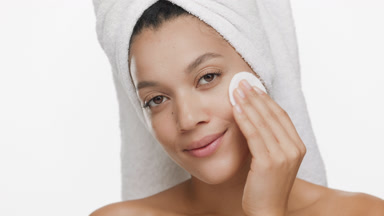 Slim woman with towel on head strokes cheek with cotton pad