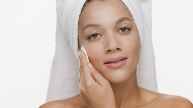Young woman with towel on hair strokes jaw line with cotton pad