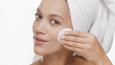 Woman with towel on head strokes face with cotton pad and smiles