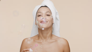 African woman with towel on hair blows soap bubbles smiling wide