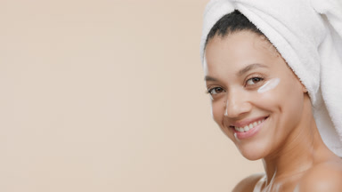 African woman with towel on hair applies cream on cheek smiling