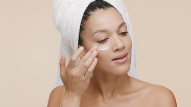 African woman with towel on head puts cream on cheek and smiles