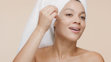 Slim woman with towel on hair drips serum from dropper on cheek