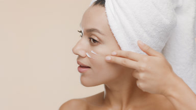 Cute woman with towel on hair applies cream on cheek and smiles