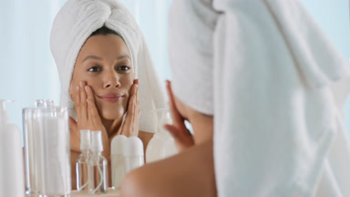 Woman with towel on hair touches cheeks in front of the mirror