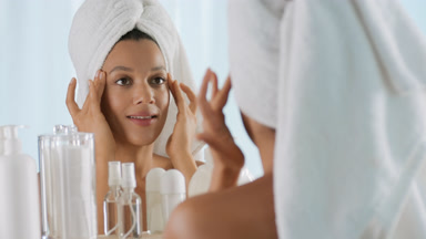 African woman with towel on head strokes face in front of mirror