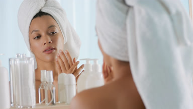 African woman with towel on hair rubs hands in front of mirror
