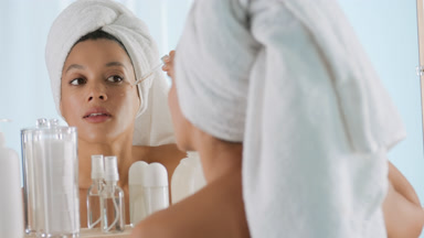 Slim woman drips serum from dropper on cheek in front of mirror