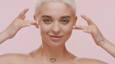 Blond woman with bare shoulders strokes face under eyes smiling