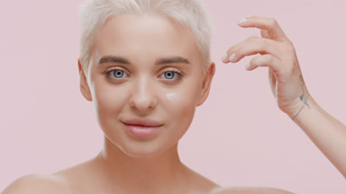 Attractive slim blond woman applies moisturizer on cheek smiling