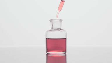Dropper drips pink oil into reagent bottle on grey background