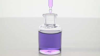 Lab dropper drips violet fluid into bottle on grey background