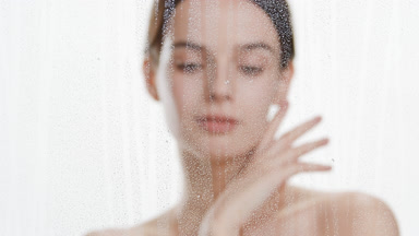 Slim woman strokes jawline behind foggy glass with rolling drops