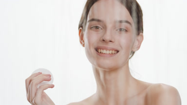 Cute slim woman strokes face using cotton pad behind foggy glass