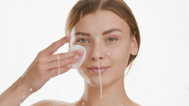 Woman strokes cheek using cotton pad behind white drops on glass