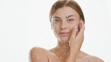 Woman strokes jawline behind glass with water drops rolling down