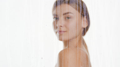 Young woman strokes shoulder and clavicles behind fogged glass