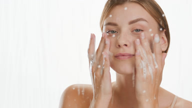Young woman applies foam on cheeks behind glass with water drops