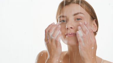 Cute young Caucasian woman puts foam on face behind wet glass