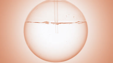 Clear liquid dropper into round flask, swirls, bubbles, beige bg