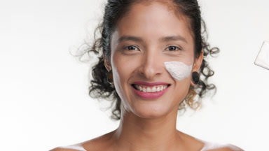 Curly-haired latina applies face clay mask with brush on white