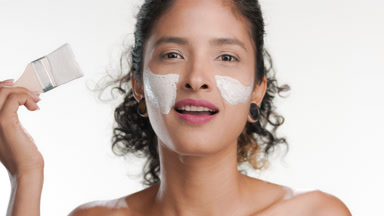 Curly-haired latina applies clay mask on cheek with brush